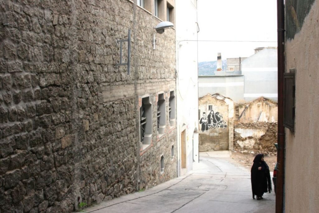 Mulher idosa caminhando por uma rua estreita de Orgosolo, vila montanhosa da Sardenha, vestida com trajes pretos tradicionais, cercada por murais históricos.