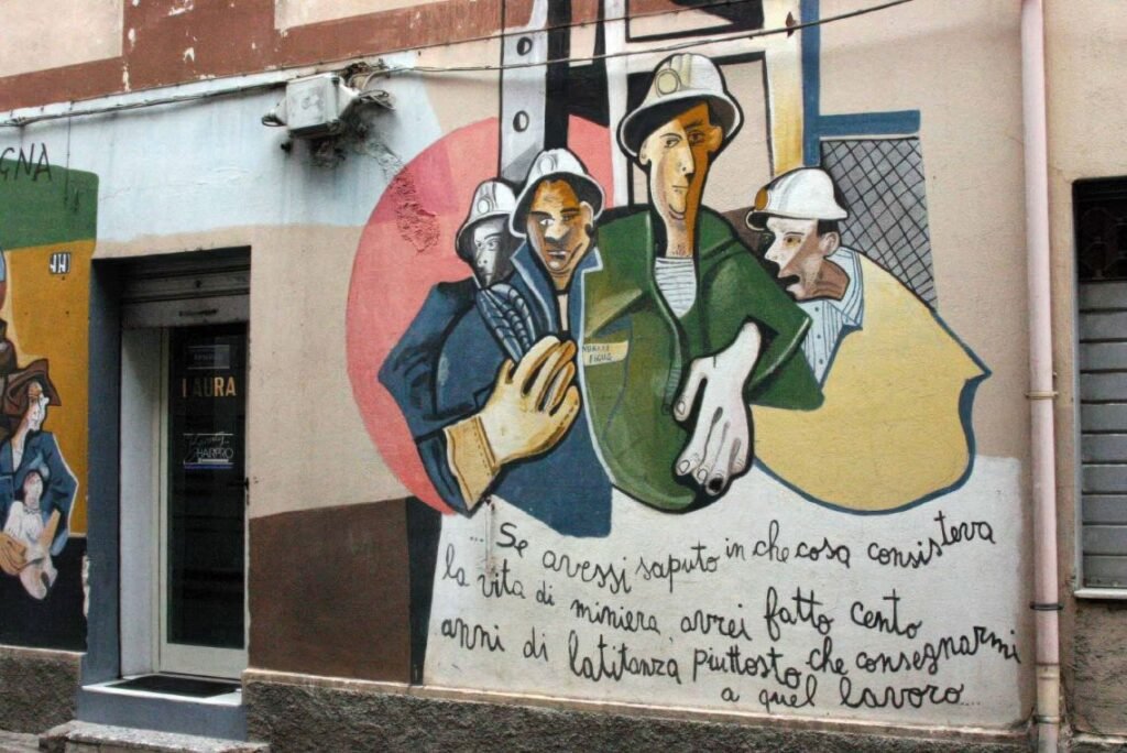 Mural representando mineiros com capacetes e luvas, ao lado de uma frase em italiano que reflete as dificuldades da mineração, localizada em um muro em Orgosolo, Sardenha.